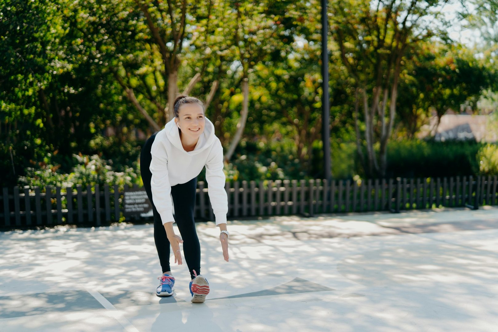 Positive female model does regular physical exercises in open air leans to raised foot in sneakers.