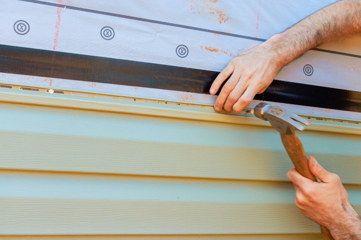 Hands working on house siding installation with hammer and protective materials during daytime