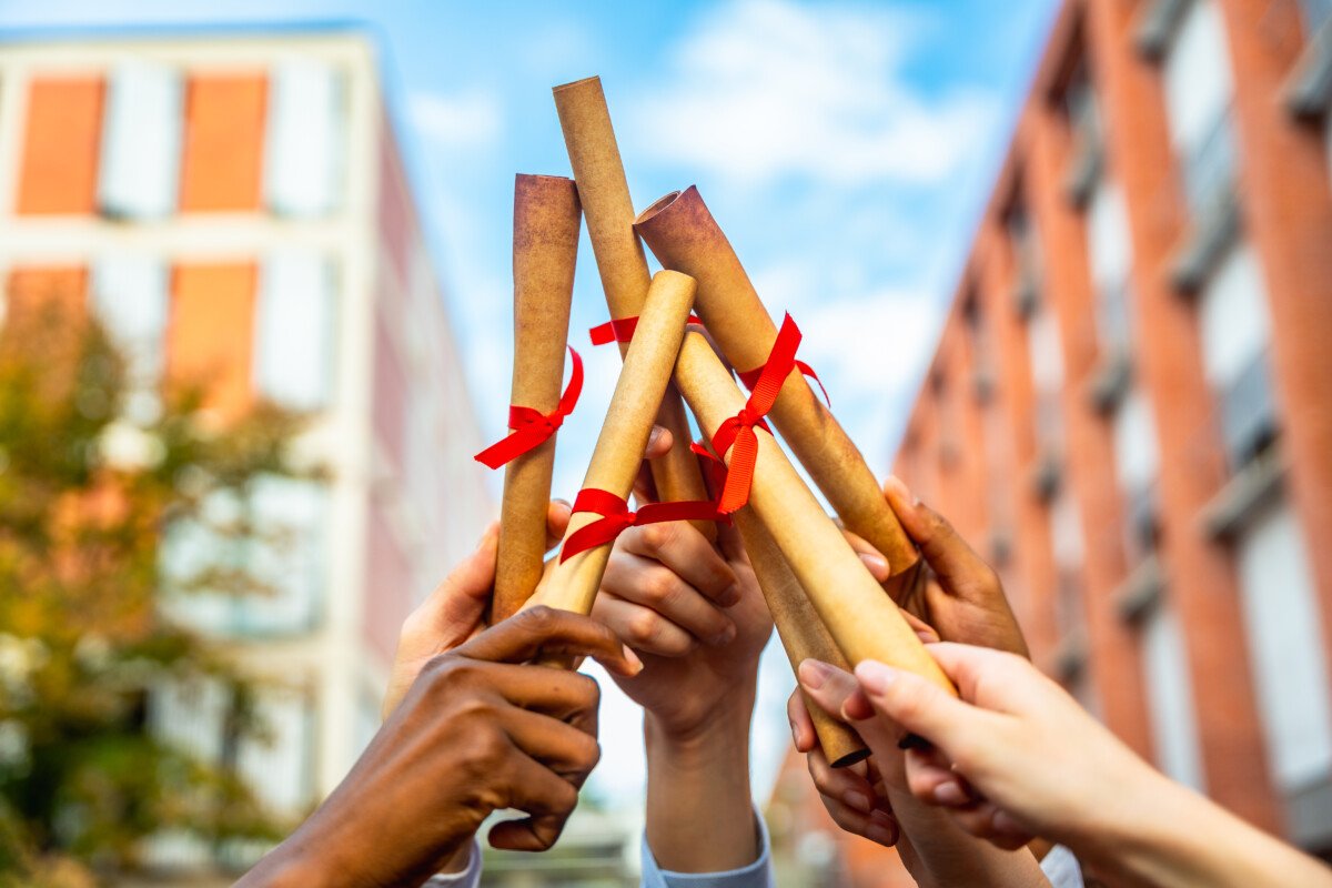 Diverse hands are holding rolled diplomas tied with red ribbons, raising them up in celebration of academic achievement and…