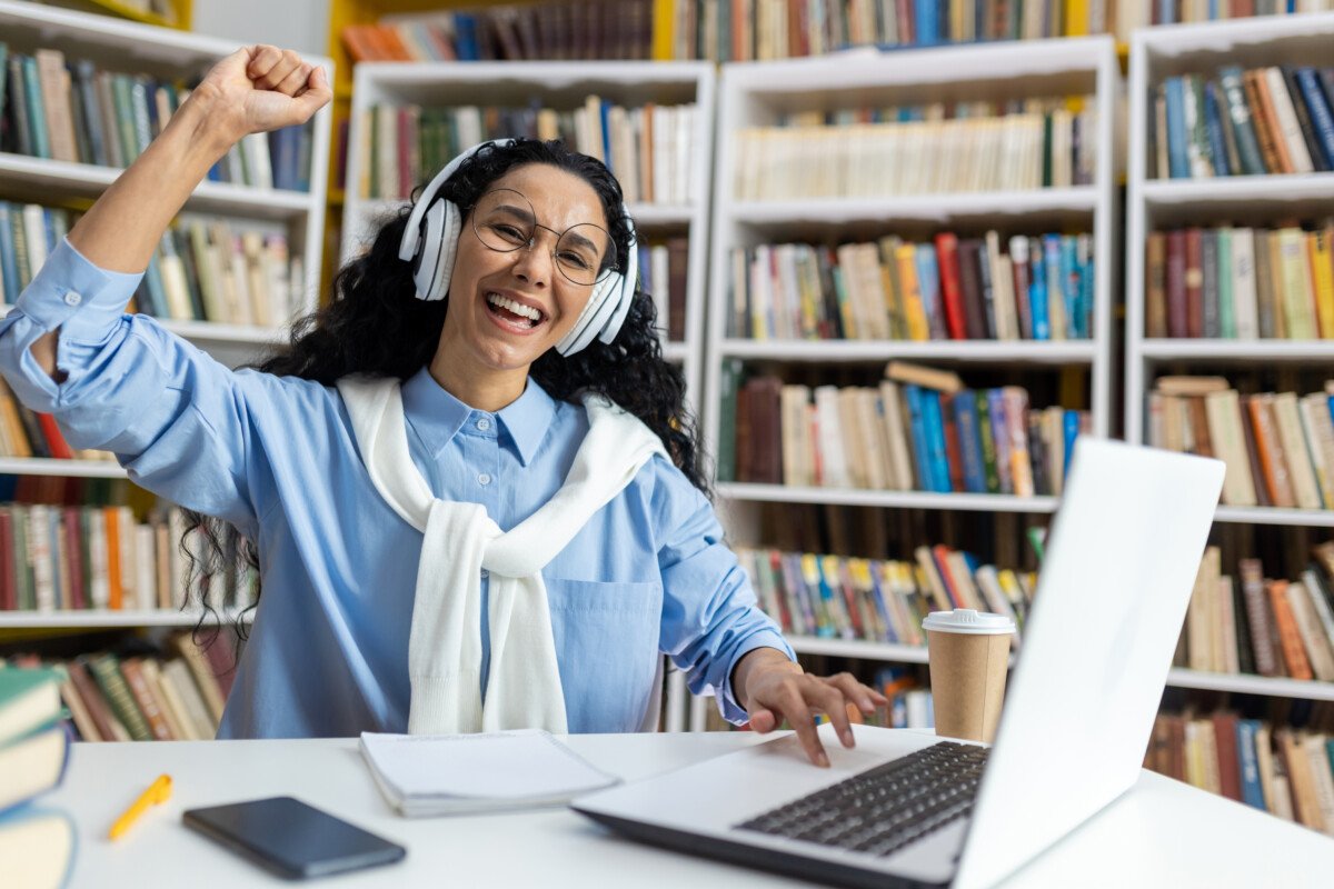 Joyful young woman with headphones cheering in front of a laptop, experiencing triumph in a library setting.