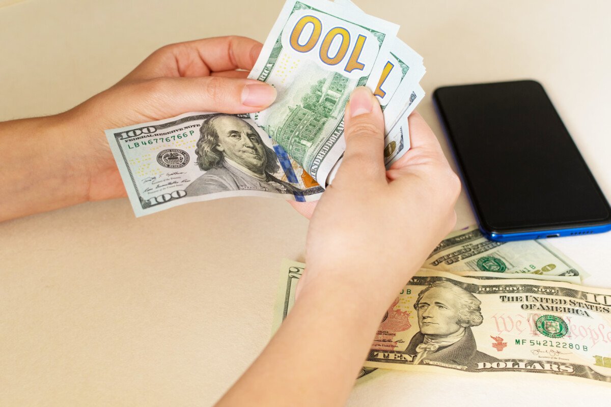 A closeup of a woman’s hands counting dollar bills, on a white desk.