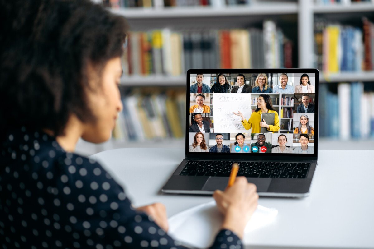 View over shoulder of an mixed race clever female student at a laptop screen with a female teacher and students. Teacher…
