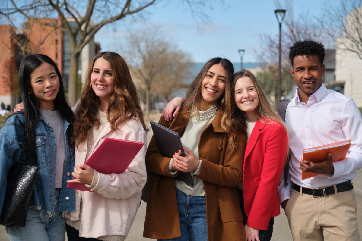 Group of cheerful multi ethnic university students smiling and holding books and a laptop on campus