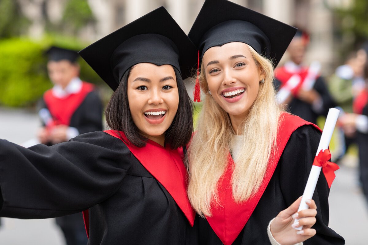 Two cheerful multiracial young ladies girlfriends in graduation clothes taking selfie, happy female students showing…
