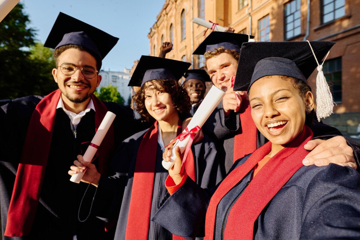Happy graduated students making selfie portrait in gowns against the university outdoors