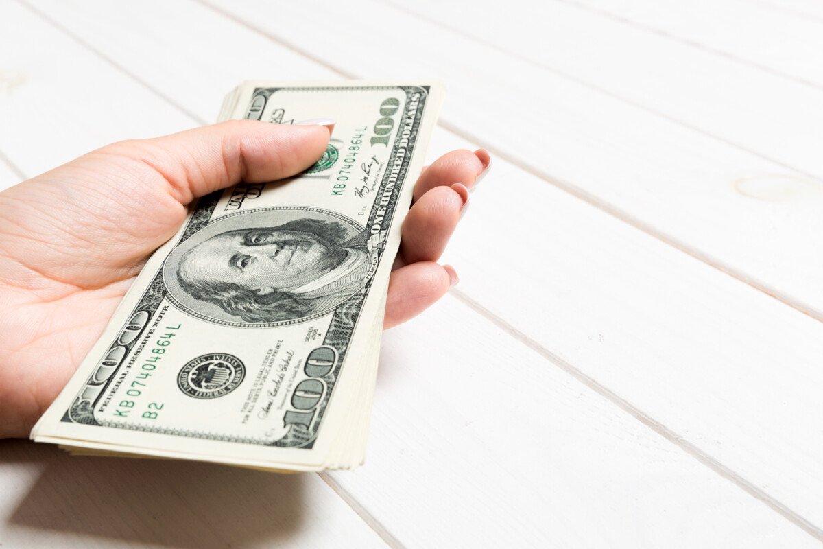 Female hand holding a bundle of money on wooden background. Perspective view of one hundred dollar banknotes. Salary…