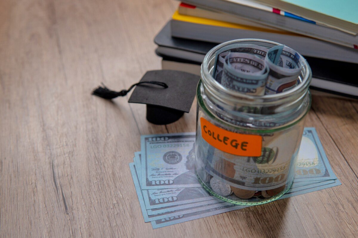A glass jar filled with coins and banknotes, labeled “COLLEGE”, a miniature graduation cap and books. Copy space for text.…