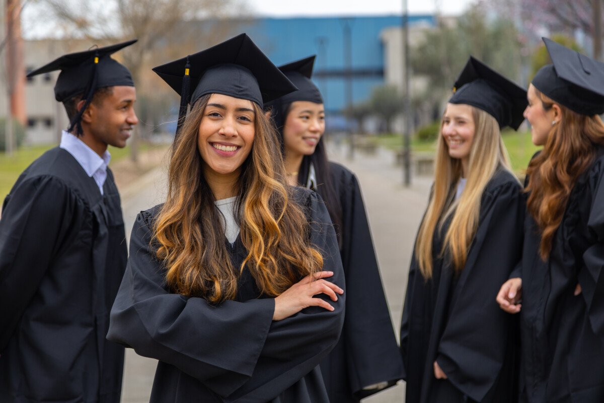Portrait of cheerful graduate student smiling with arms crossed while standing with her colleagues outdoors after the…