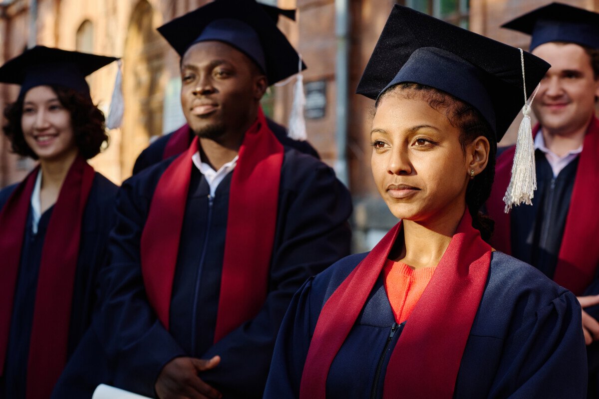 Group of students in graduation gowns listening to speaker while standing outdoors