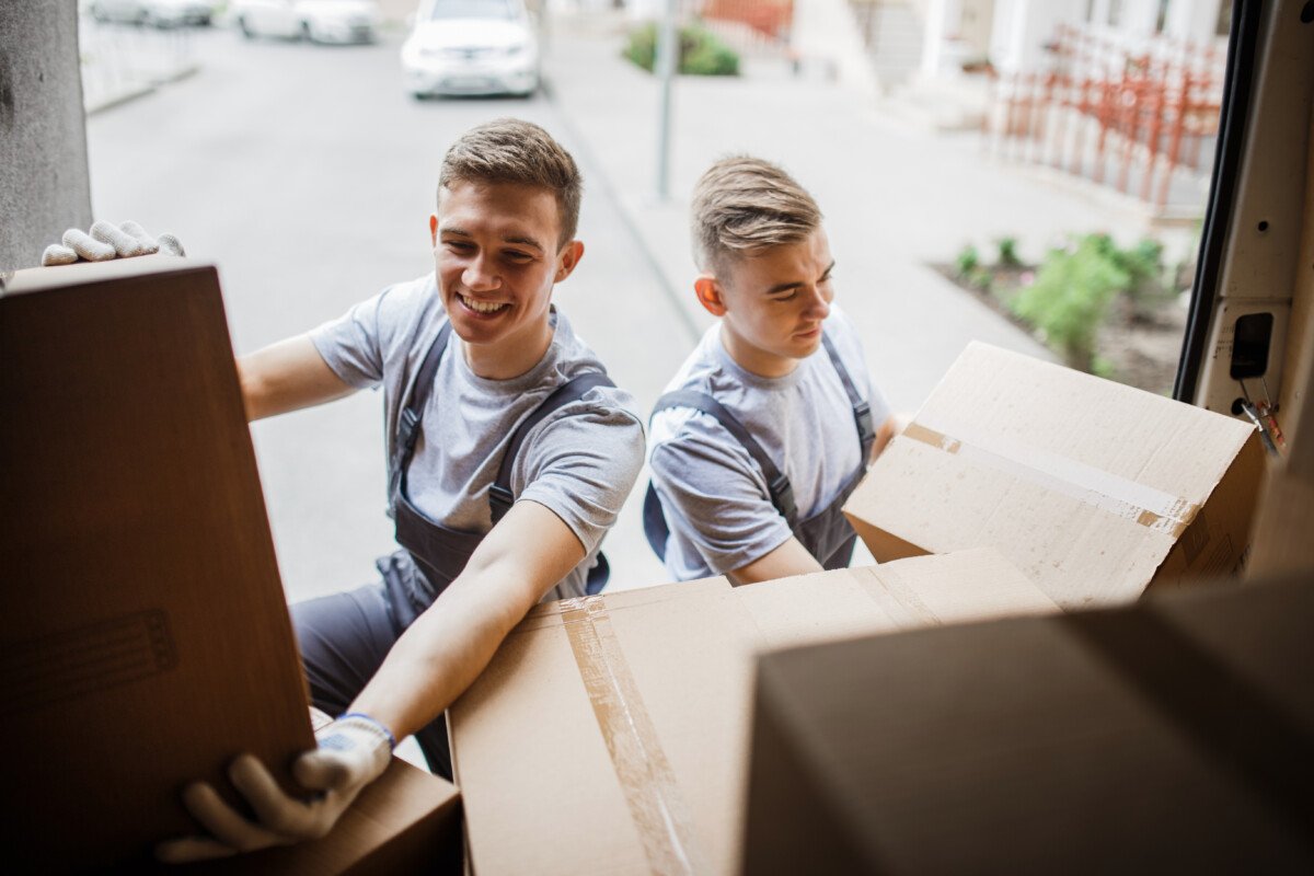 Two young handsome smiling movers wearing uniforms are unloading the van full of boxes. House move, mover service.