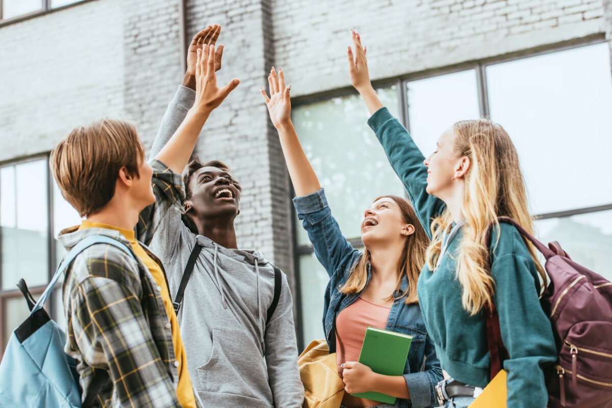 Selective focus of positive multicultural teenagers with backpacks and raised hands outdoors