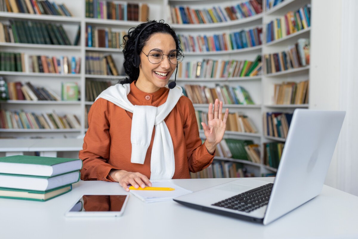 Joyful curly Hispanic girl sitting at a table in front of a laptop in a headset looking at the camera and waving at the…