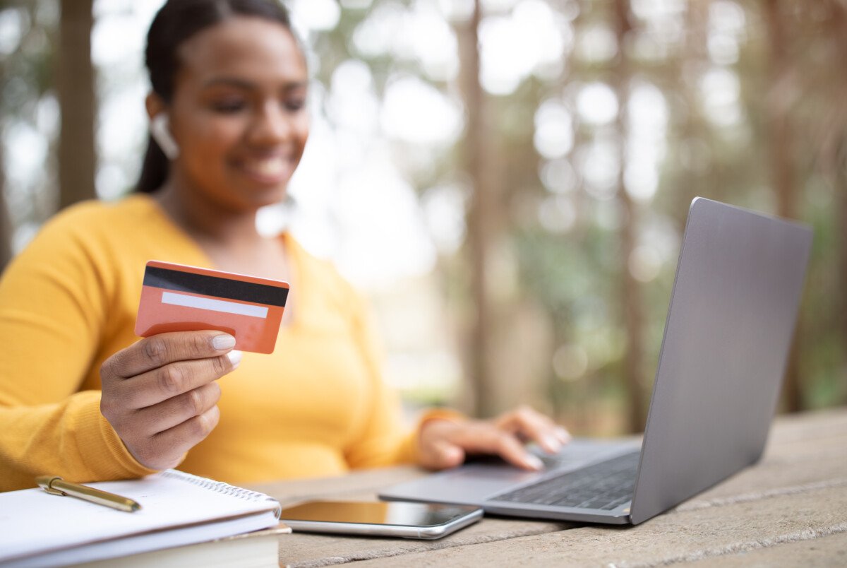 Online payment. Brazilian lady with laptop and credit card sitting outdoors in park, smiling female enjoying internet…