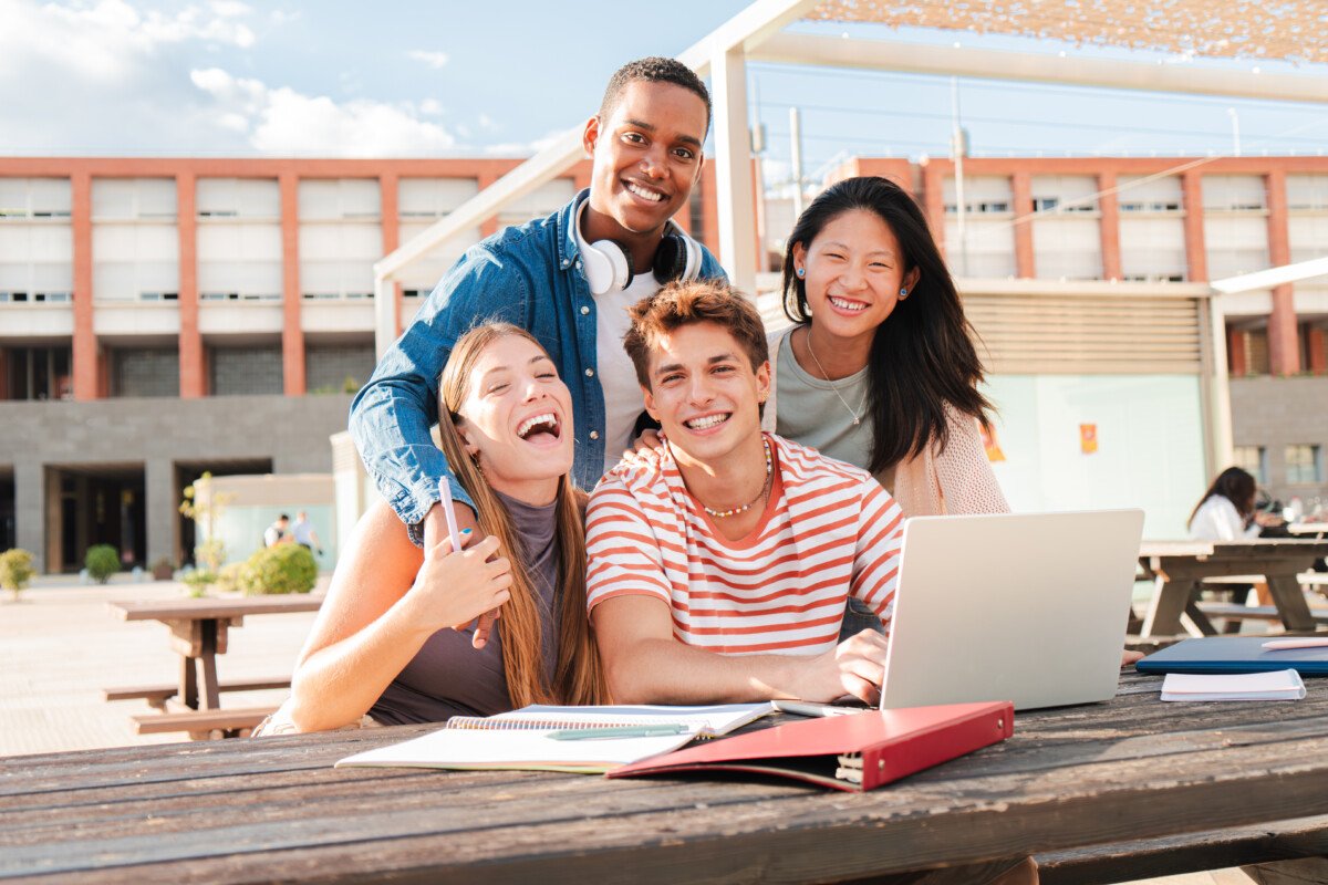 Diverse group of male and female students, smiling and looking at camera while studying together using a laptop on a…