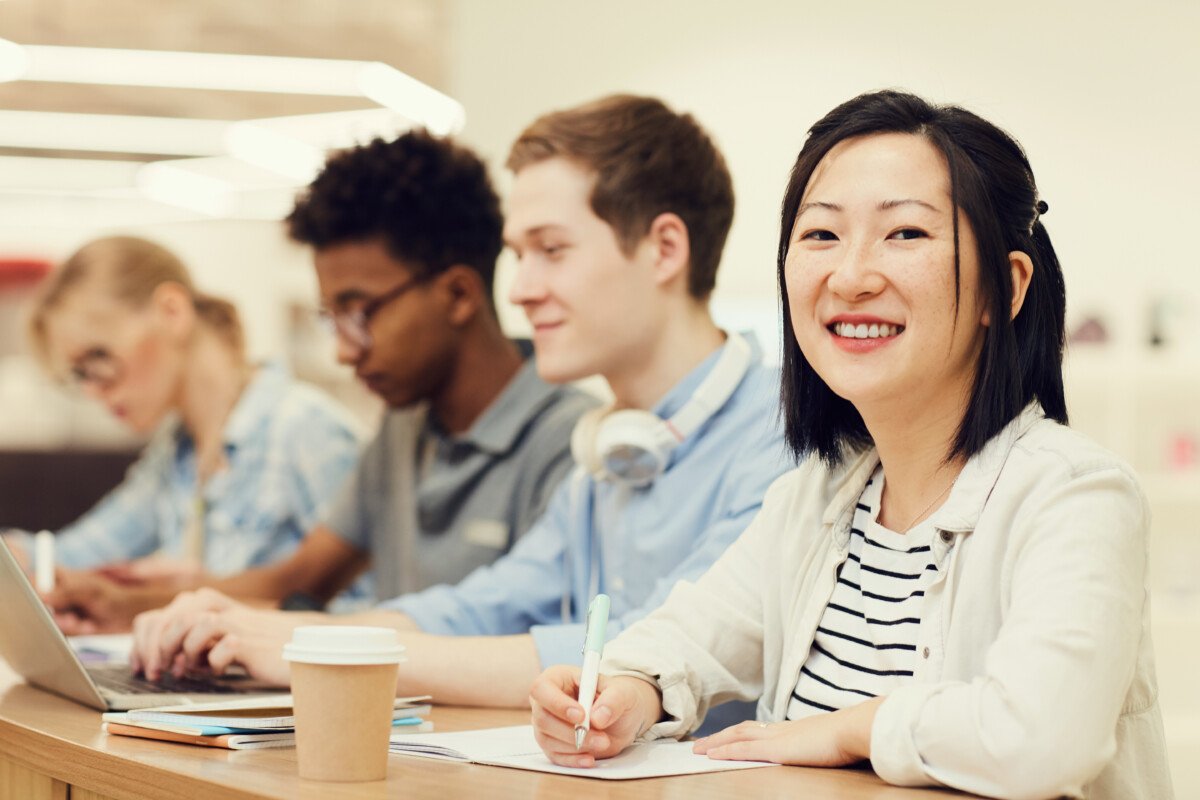 Portrait of smiling young Asian female student with short hair sitting at table with groupmates in lecture room and…