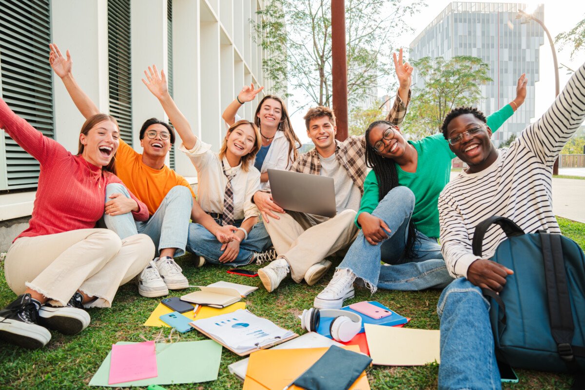 Portrait of a big group of happy teenage students sitting at university campus lawn, gesturing and celebrating together a…