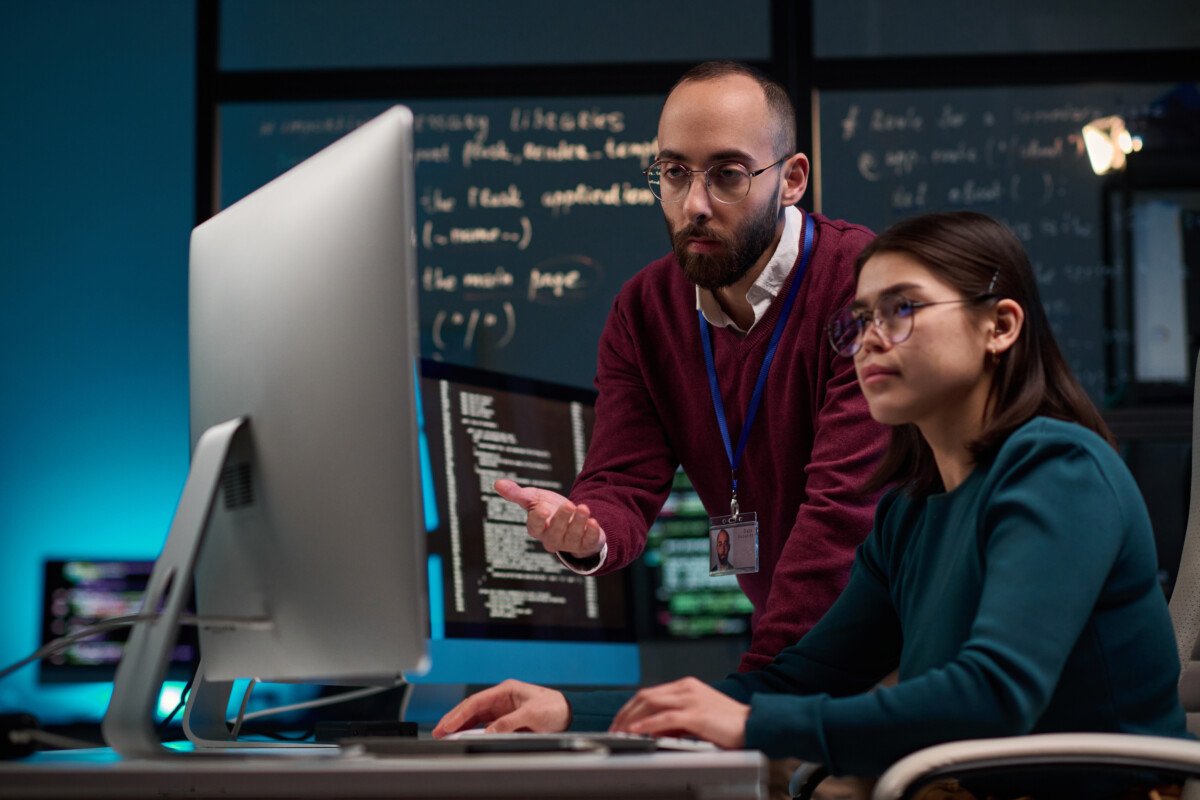 Portrait of bearded adult man as IT development supervisor advising young trainee in office and pointing at computer screen…