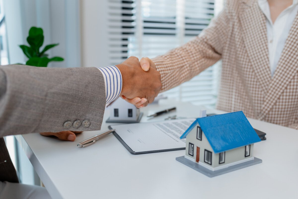 Two people in business attire finalize a real estate deal, shaking hands across a desk with house models, keys, and a…