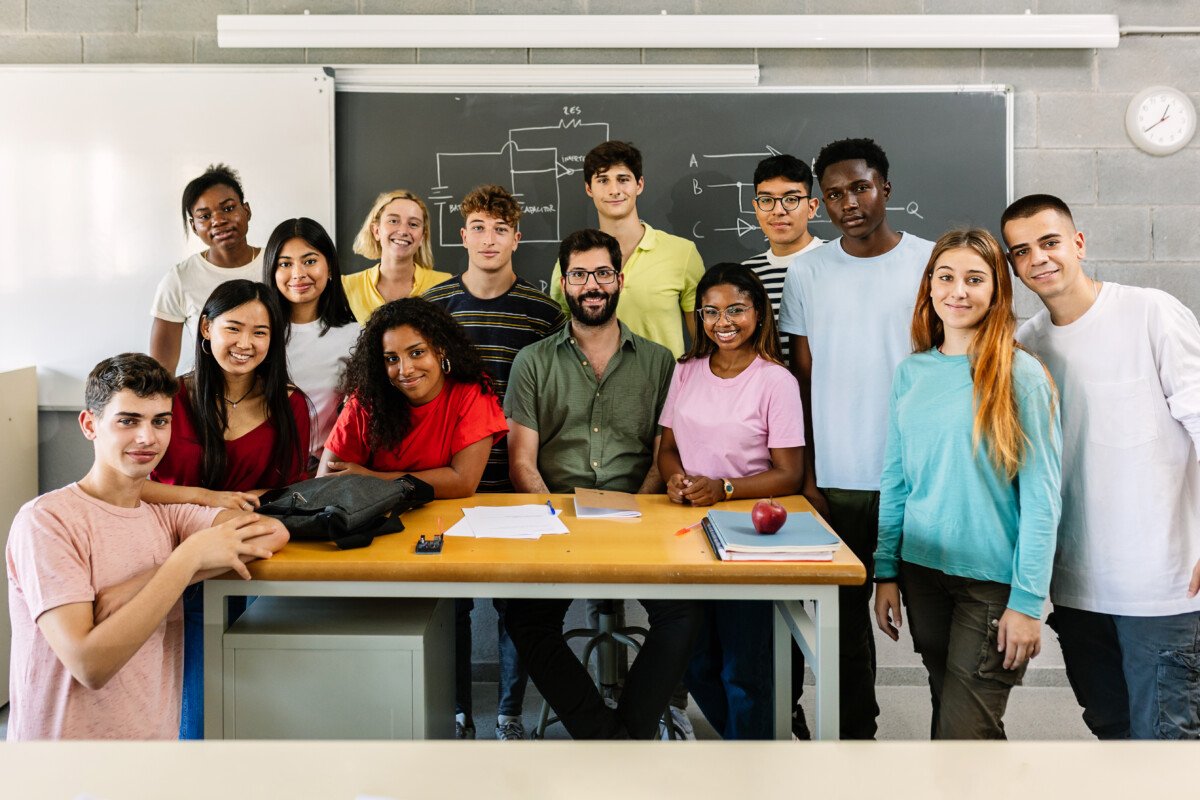 Large group portrait of millennial diverse students with male teacher smiling at camera standing together in classroom…