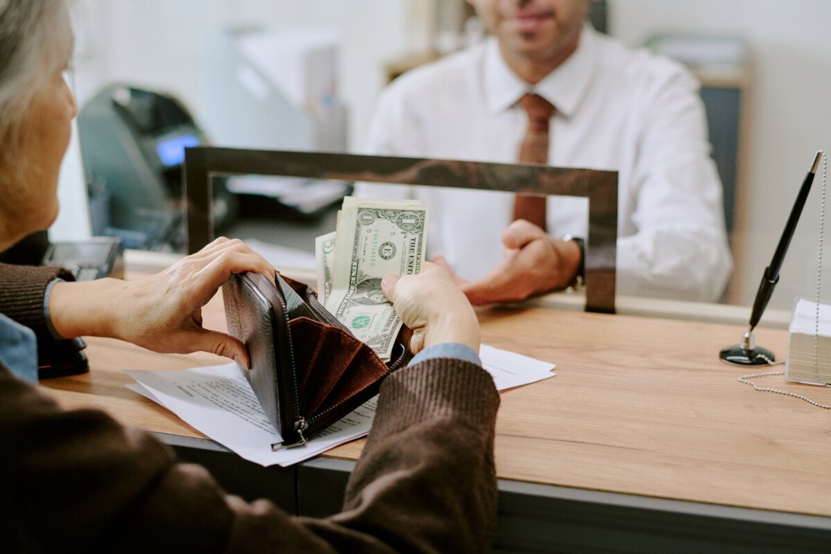 Senior person handing money to bank employee across counter in professional setting, financial transaction taking place in…