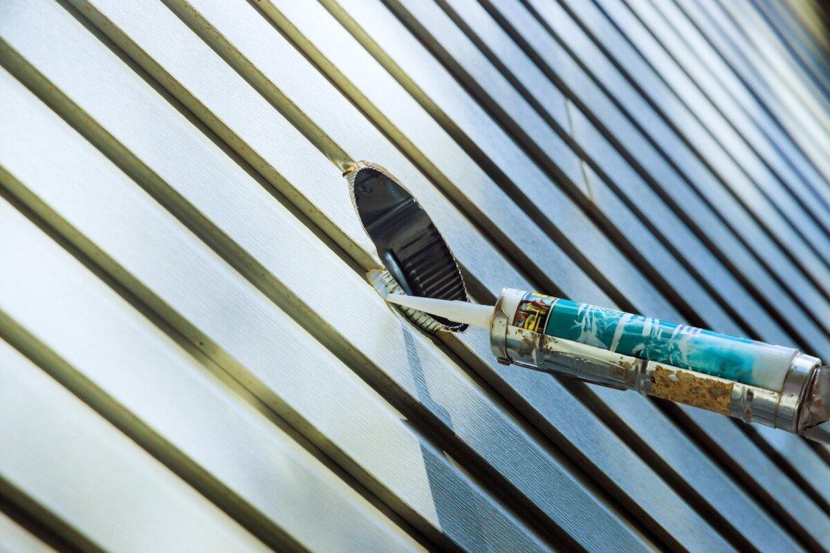 Repairing exterior siding with a putty knife and caulk gun in a sunny outdoor setting