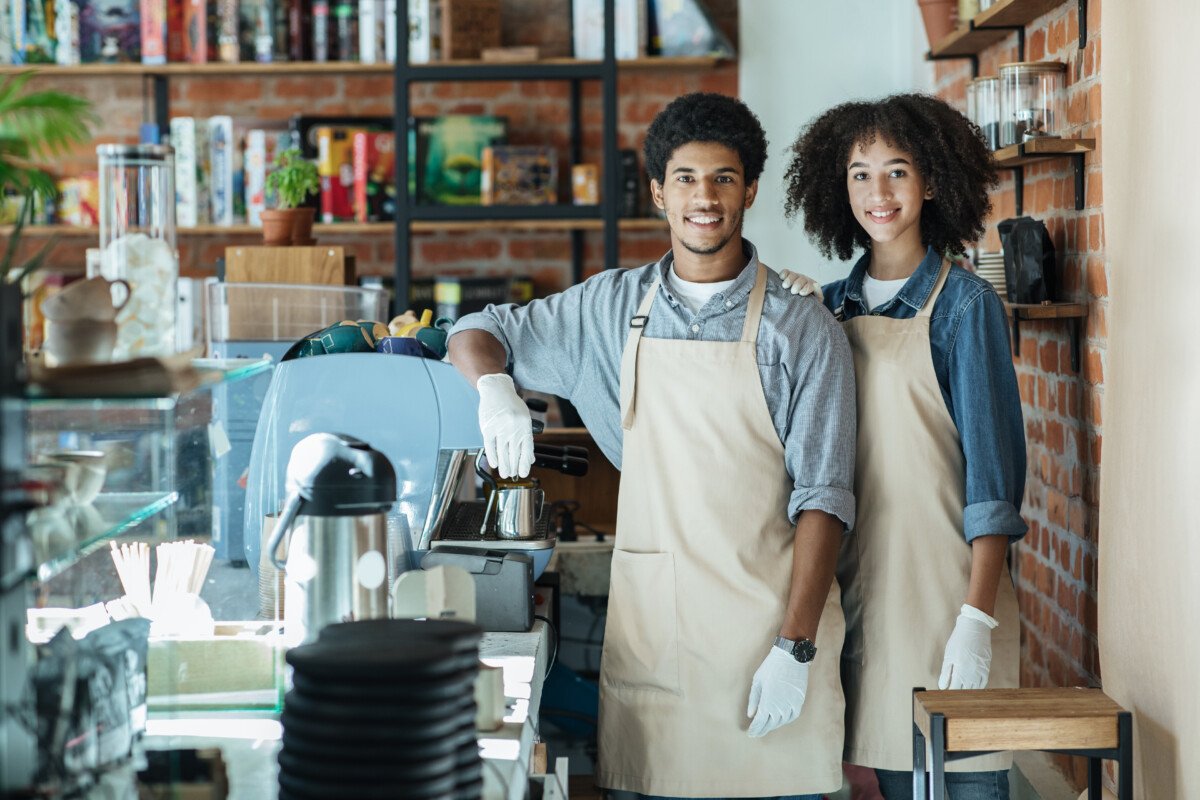 Meeting clients in cafe after covid-19 lockdown. Smiling african american guy and female in aprons and white gloves with…