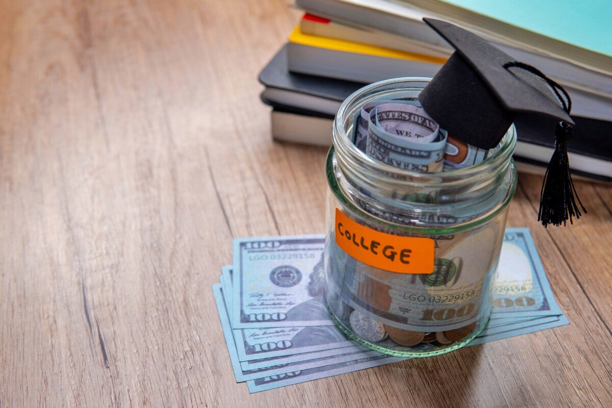 A glass jar filled with coins and banknotes, labeled “COLLEGE”, a miniature graduation cap and books. Copy space for text.…