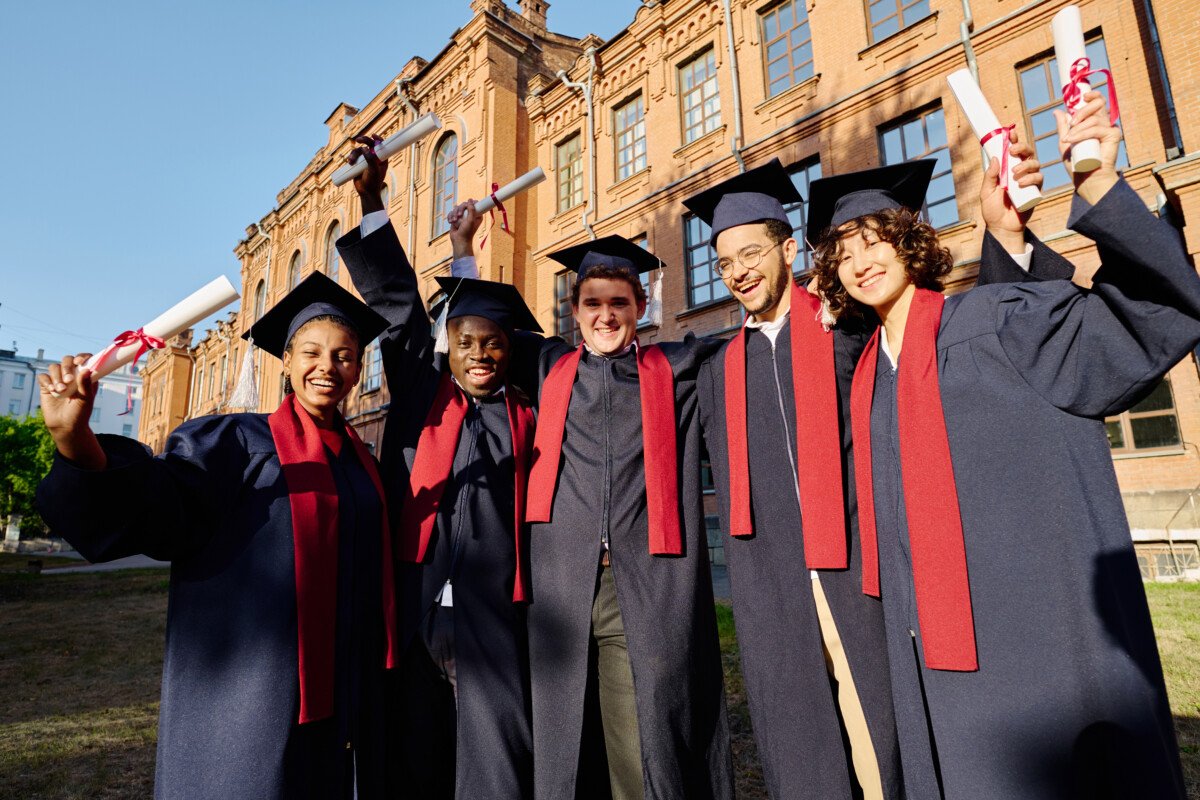 Group of happy students in gowns standing with diplomas outdoors near the university