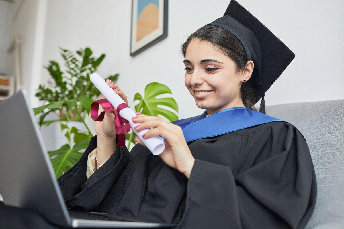 Portrait of young woman wearing graduation gown and using laptop while speaking by video chat indoors