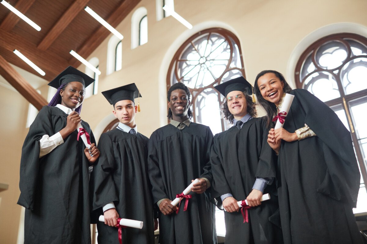Low angle view at multi-ethnic group of young people wearing graduation robes and looking at camera smiling happily while…