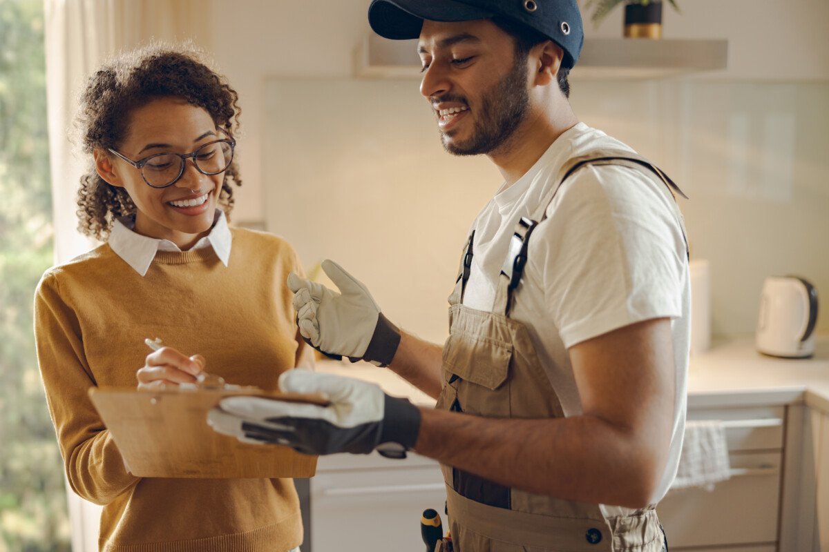 Smilng young woman signing document while communicating with handyman at the kitchen