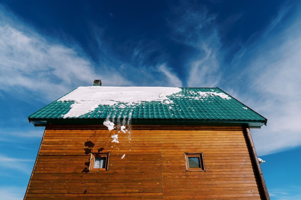Snow falls from the green tiled roof of a wooden cottage against the blue sky. High quality photo