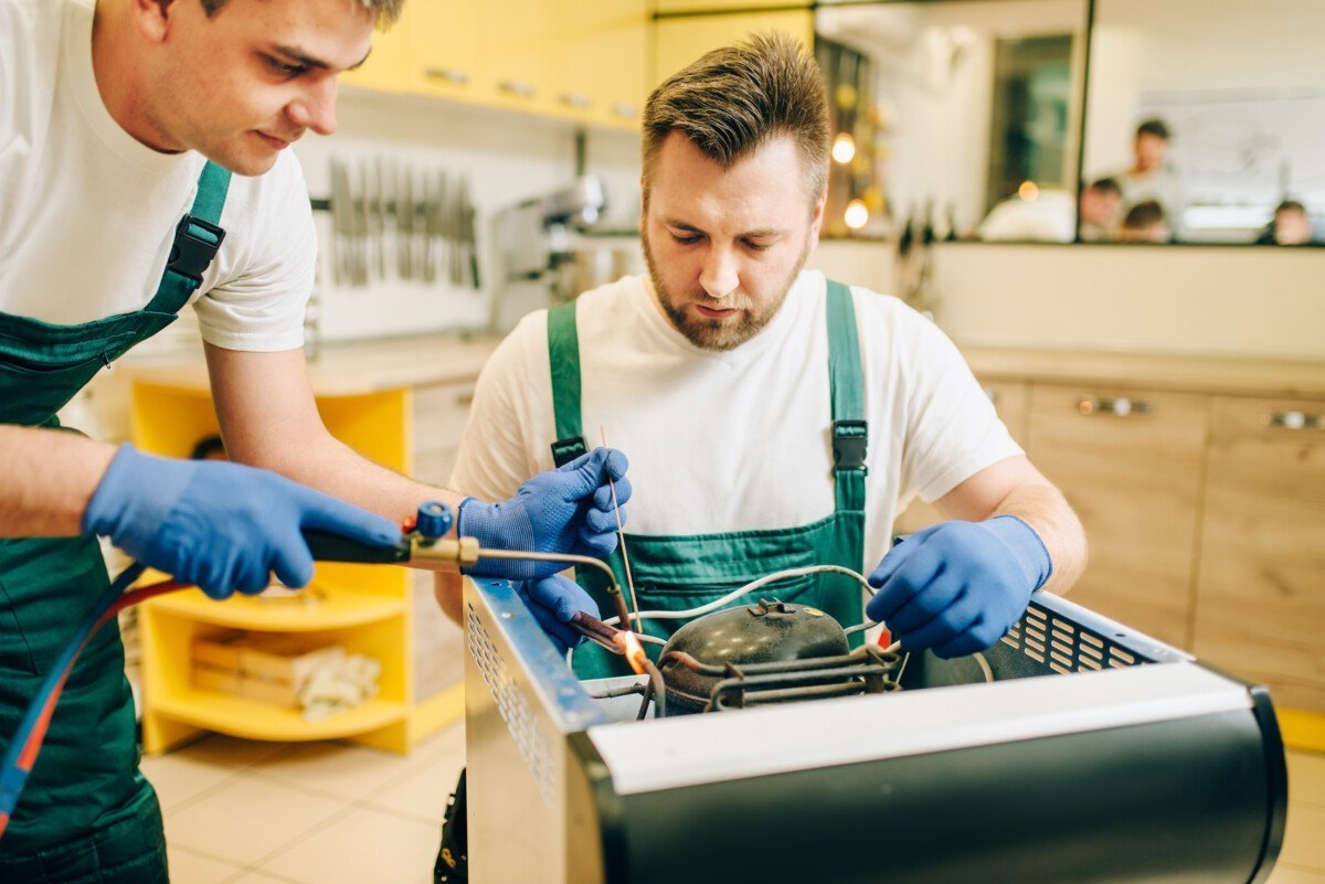 Workers with the burner repairs refrigerator compressor at home. Repairing of fridge occupation, professional service