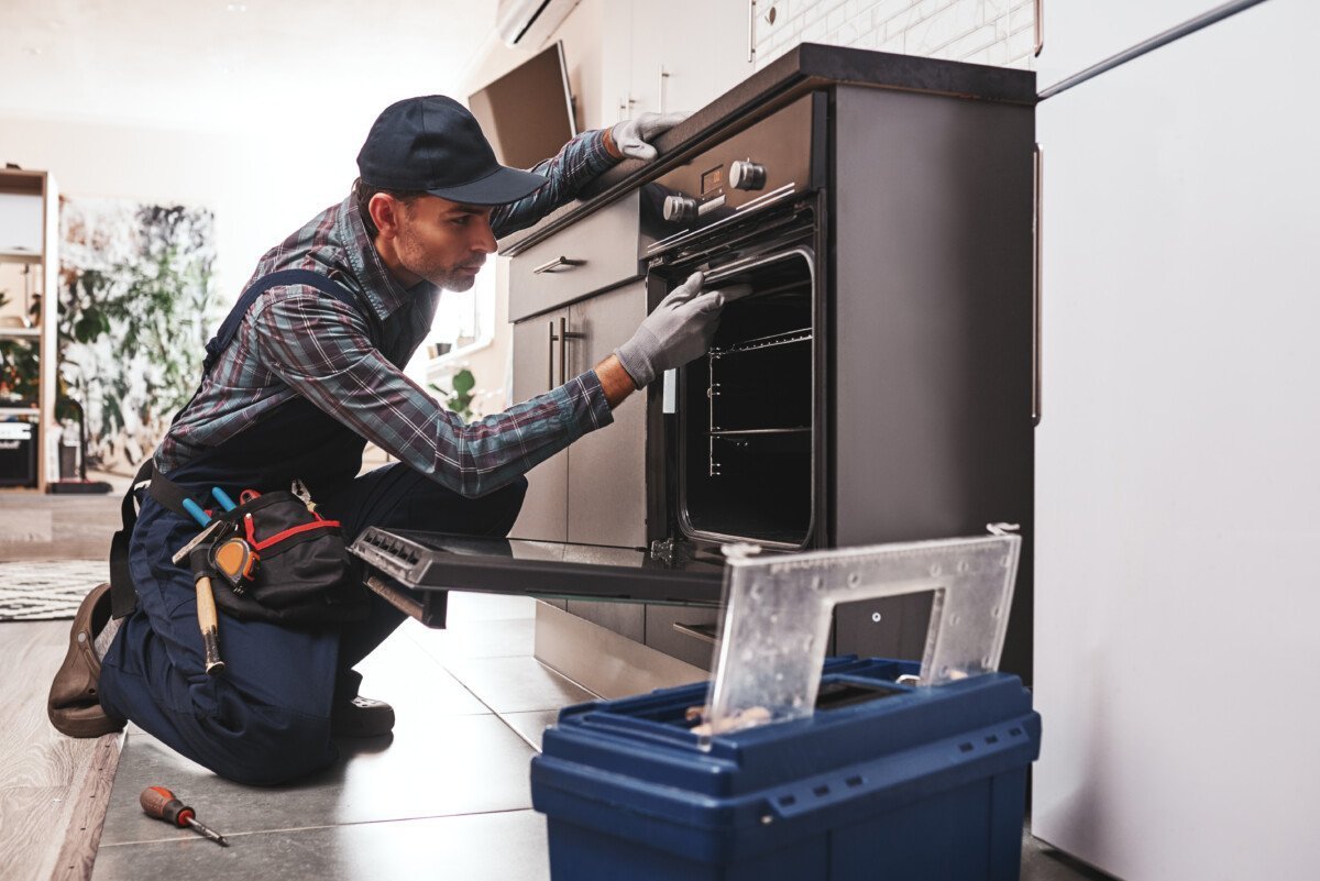 Don’t delay with repair. Close-up of repairman examining oven with screwdriver in kitchen with tool case