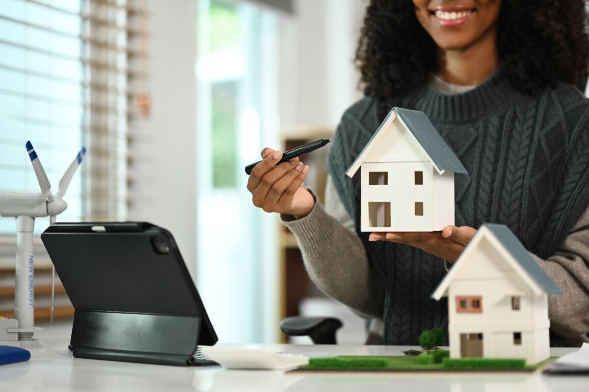 Smiling African American woman pointing at miniature model of a house. Real estate appraisal and property concept.