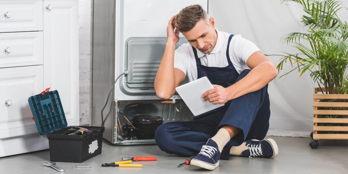 thoughtful adult repairman sitting on floor and using digital tablet while repairing refrigerator