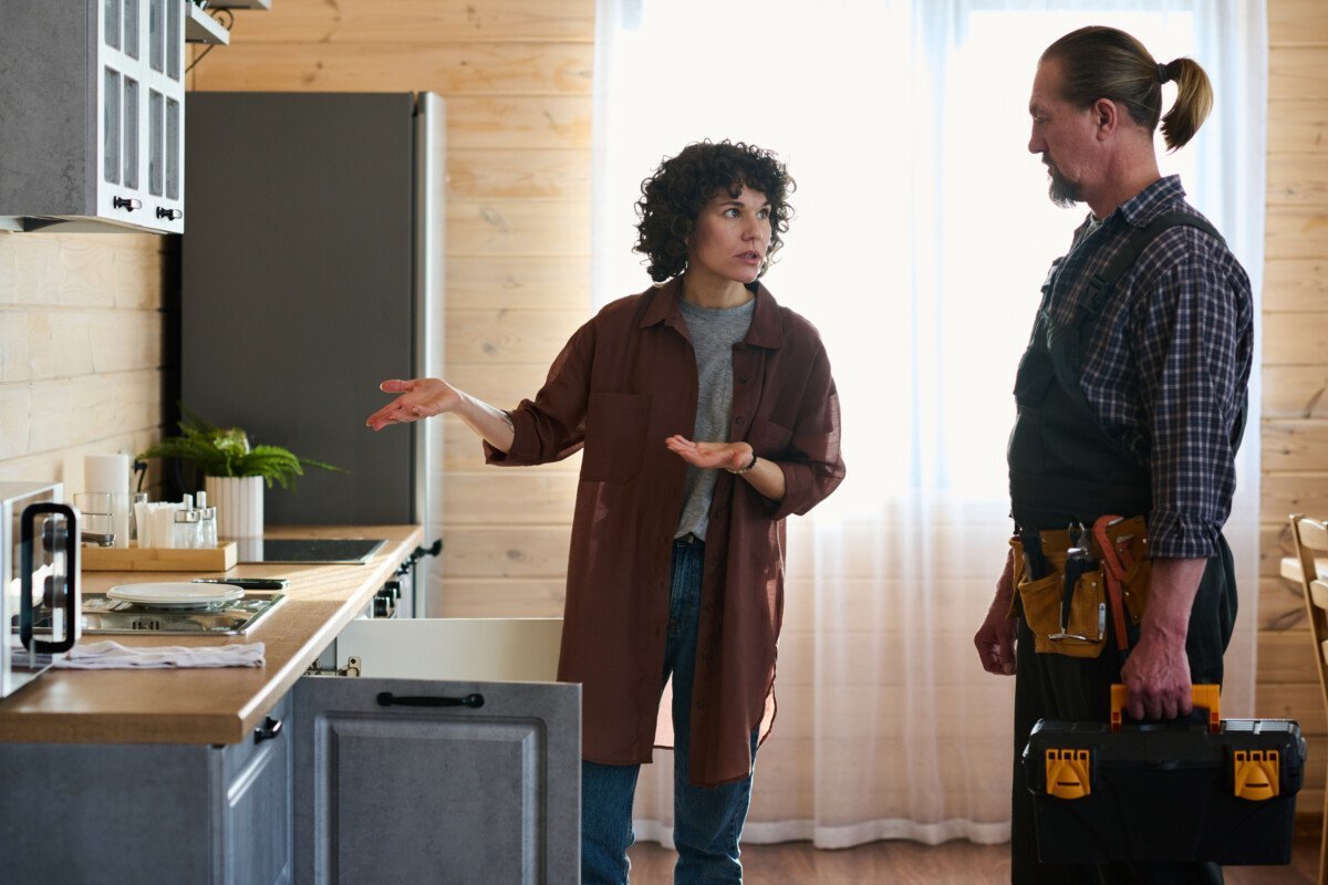 Young worried housewife looking at plumber and explaining him problem with pipe or faucet while standing by kitchen counter…